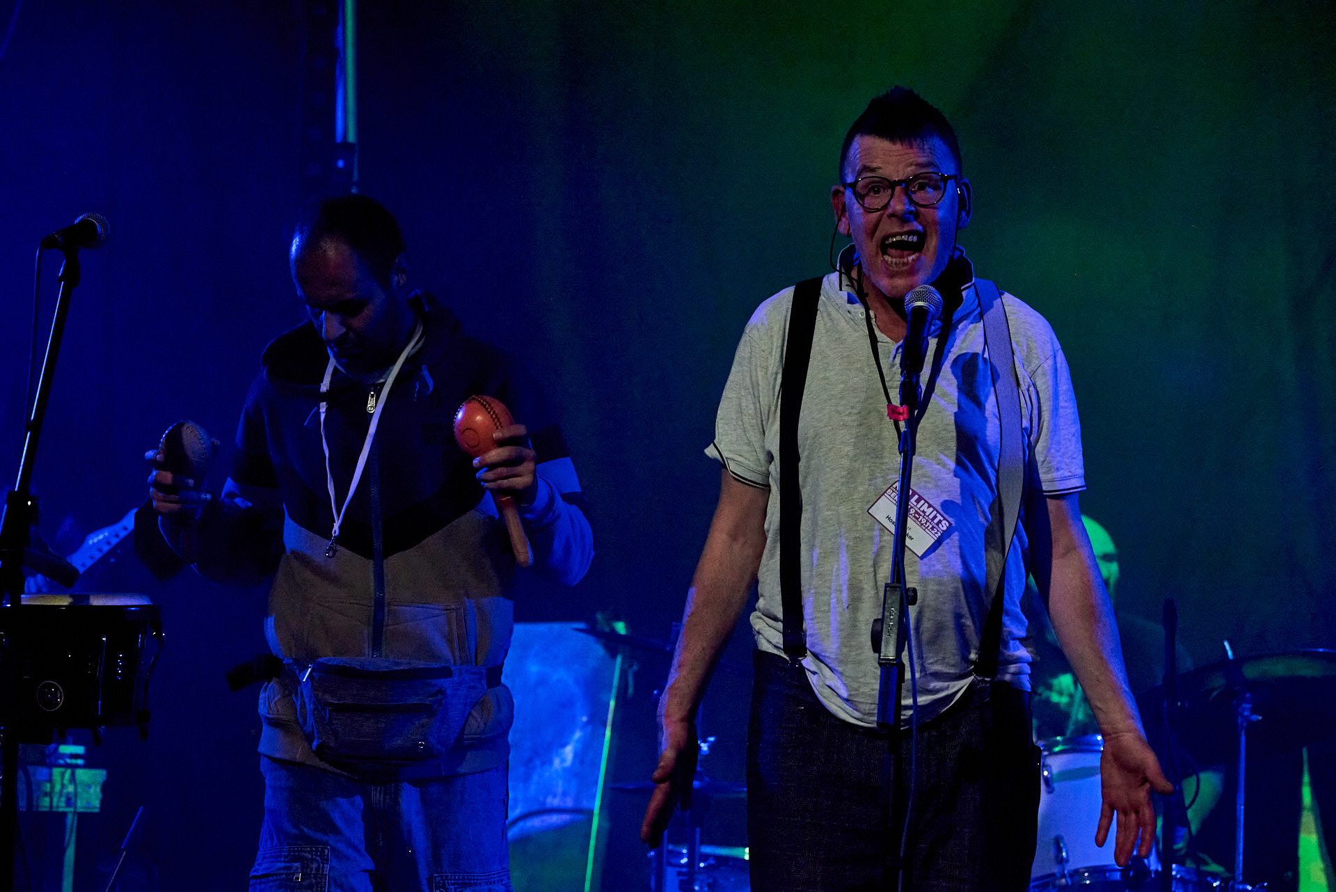 Sänger Hoss mit weit geöffnetem Mund singend auf der Bühne vor seinen Bandkollegen mit Rassel im Hintergrund.