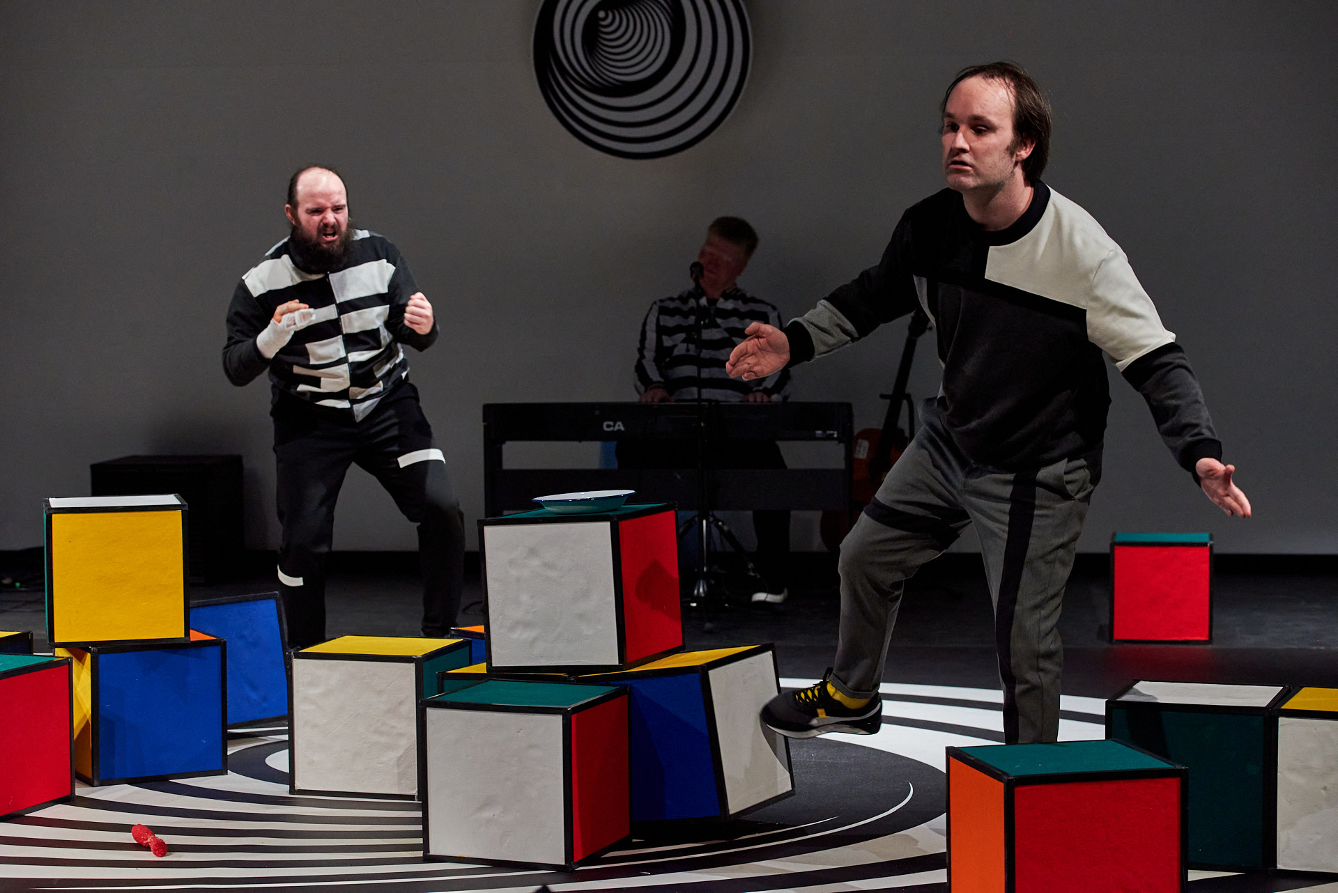 Auf dem Bild sieht man die Hauptdarsteller Max Edgar Freitag und Frank Schulz tanzend. Im Hintergrund sieht man die aufgestellten, bunten Zauberwürfel und einen weiteren Performer an einem E-Piano. 