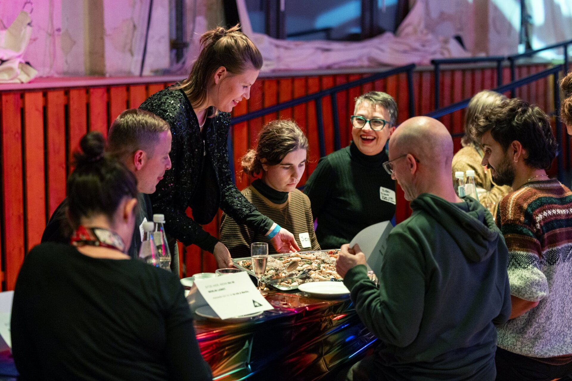 Eine Gruppe Menschen sitzt um einen langen Tisch versammelt. Sie unterhalten sich, diskutieren miteinander und lachen, während der Tisch mit Essen gedeckt wird.