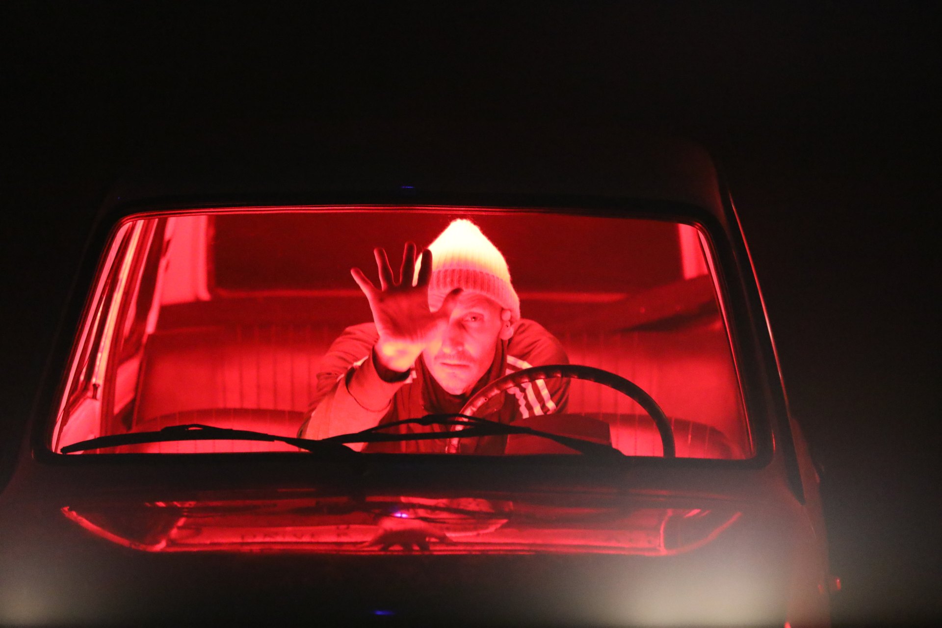 Marc Brew sitting inside a car illuminated by red light, reaching out with one hand toward the windshield.