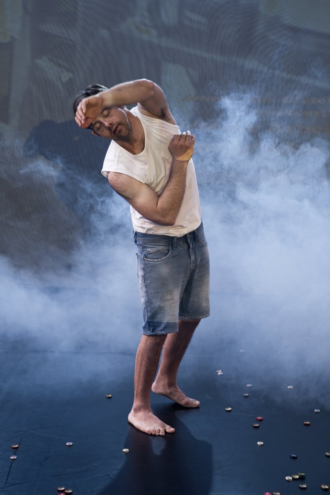 Oskar Spatz im weißen T-Shirt und Jeans-Shorts. Einem Arm über den Kopf und dem anderen angewinkelt. Um ihn herum ist künstlicher Nebel, und auf dem Boden liegen Kronkorken. 